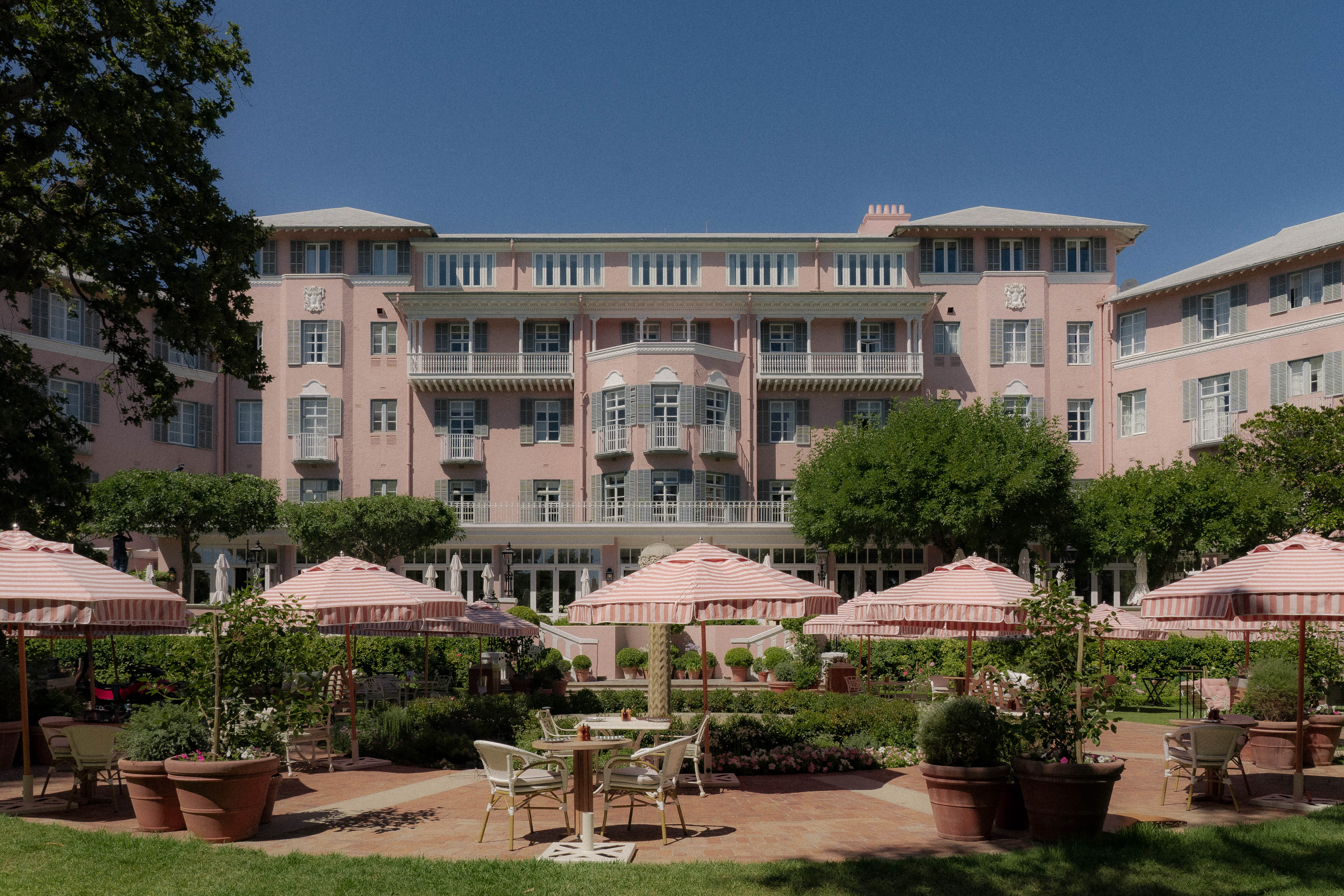 The exterior of a pale pink-tinted, neo-colonial-style hotel, with stripy parasols in pink and white stripes, romantic tables, large ceramic planters, and lots of greenery spread in the sun.