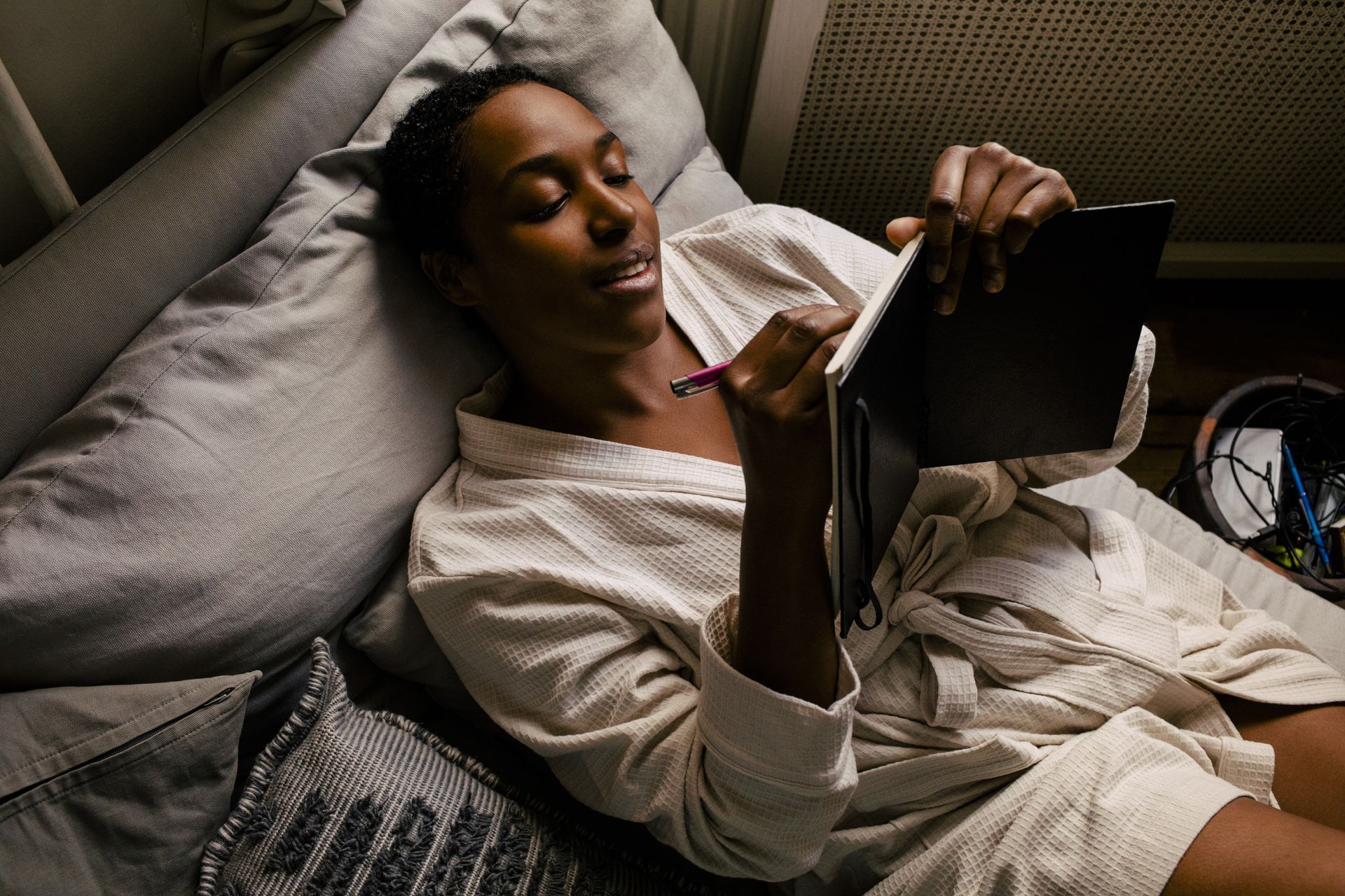 A woman lies in bed wearing a dressing gown as she writes in a journal.