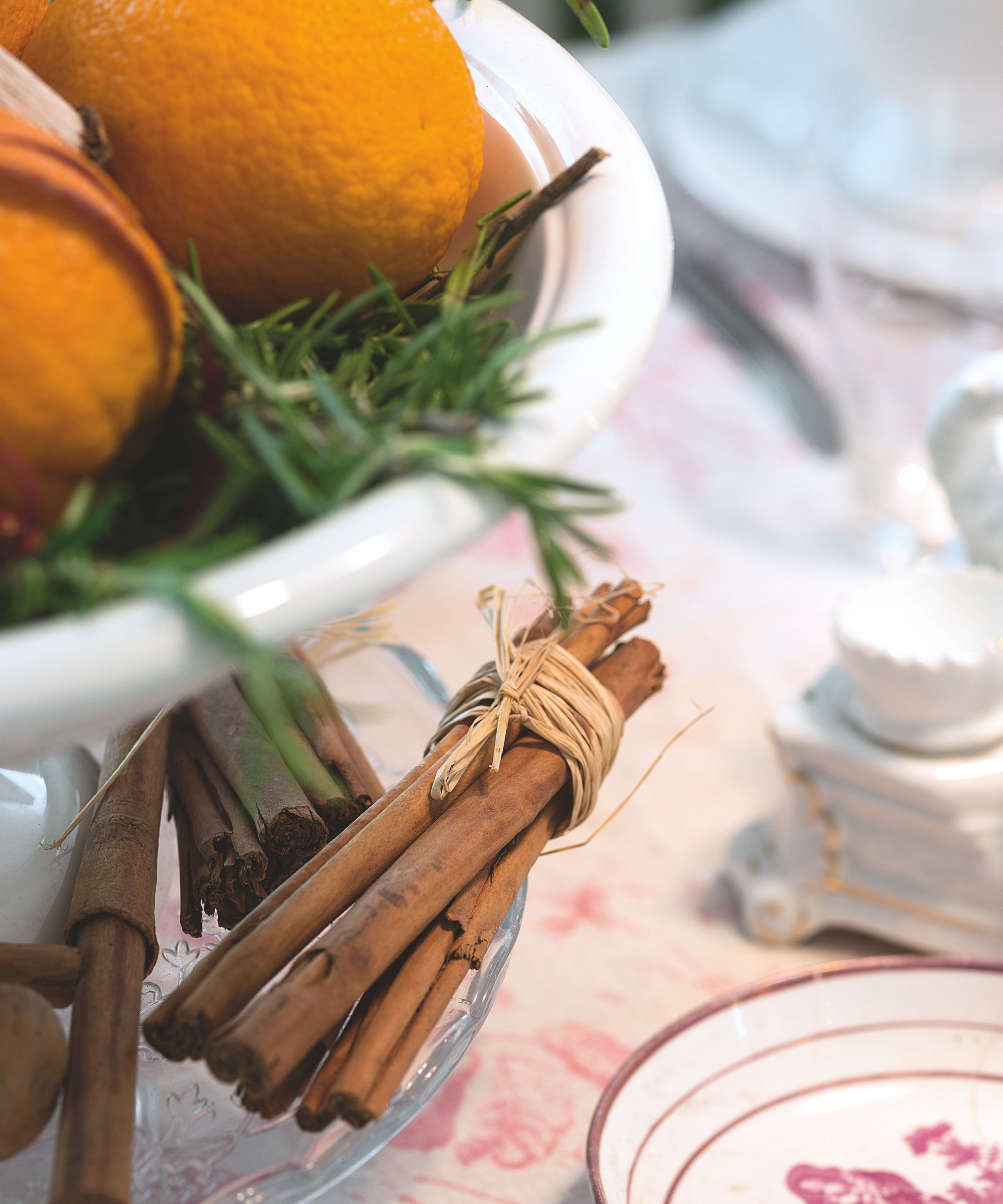In the left is a large white fruit bowl filled with oranges and fir needles, on a large glass plate with bundles of cinnamon sticks tied with string. To the right is the corner of the plate, and in the blurred background a patterned tablecloth and tableware are seen.