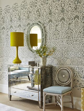 Mirrored chest of drawer and yellow table lamp and side chair in bedroom with patterned wallpaper and wall mirror