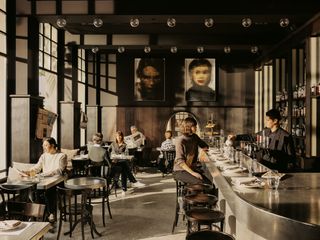 Restaurant ara, with metal tables and wooden chairs arranged throughout the room. To the right, the is a metal bar area, with dark wood bar chairs.