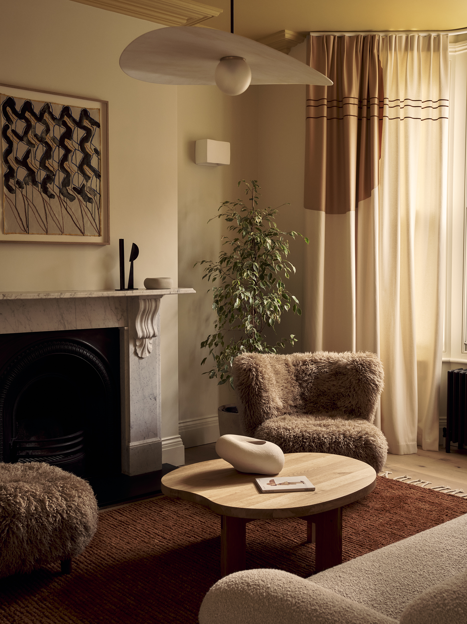 neutral living room with cream walls, fuzzy brown armchairs, terracotta rug, timber round coffee table, cream sofa, with marble fireplace with artwork on top and pendant light