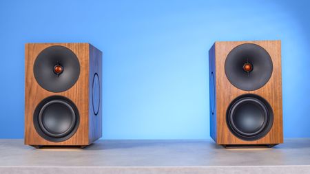 The Cambridge Audio L/R X speakers in walnut color, positioned on a stone surface with a blue background