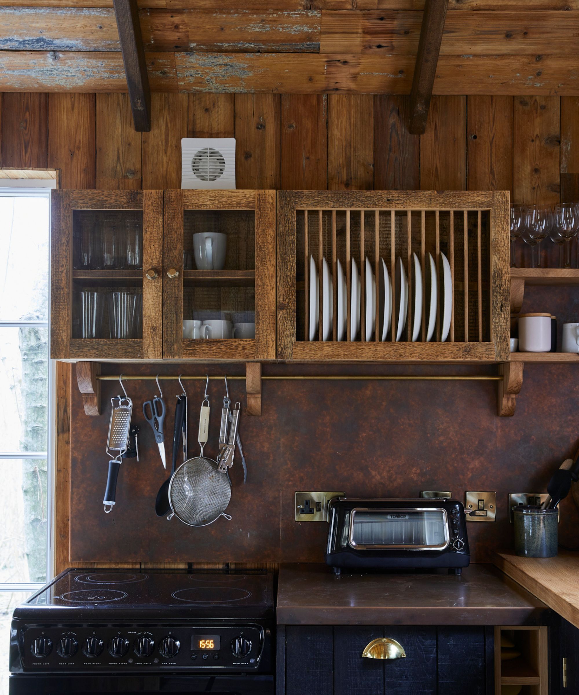 A charming rustic kitchen inside a timber cabin, complete with reclaimed wood cabinetry, a vintage-style dish rack, and bronze-toned splashback. Hanging utensils and retro appliances add to the characterful, handcrafted aesthetic, ideal for a woodland retreat.