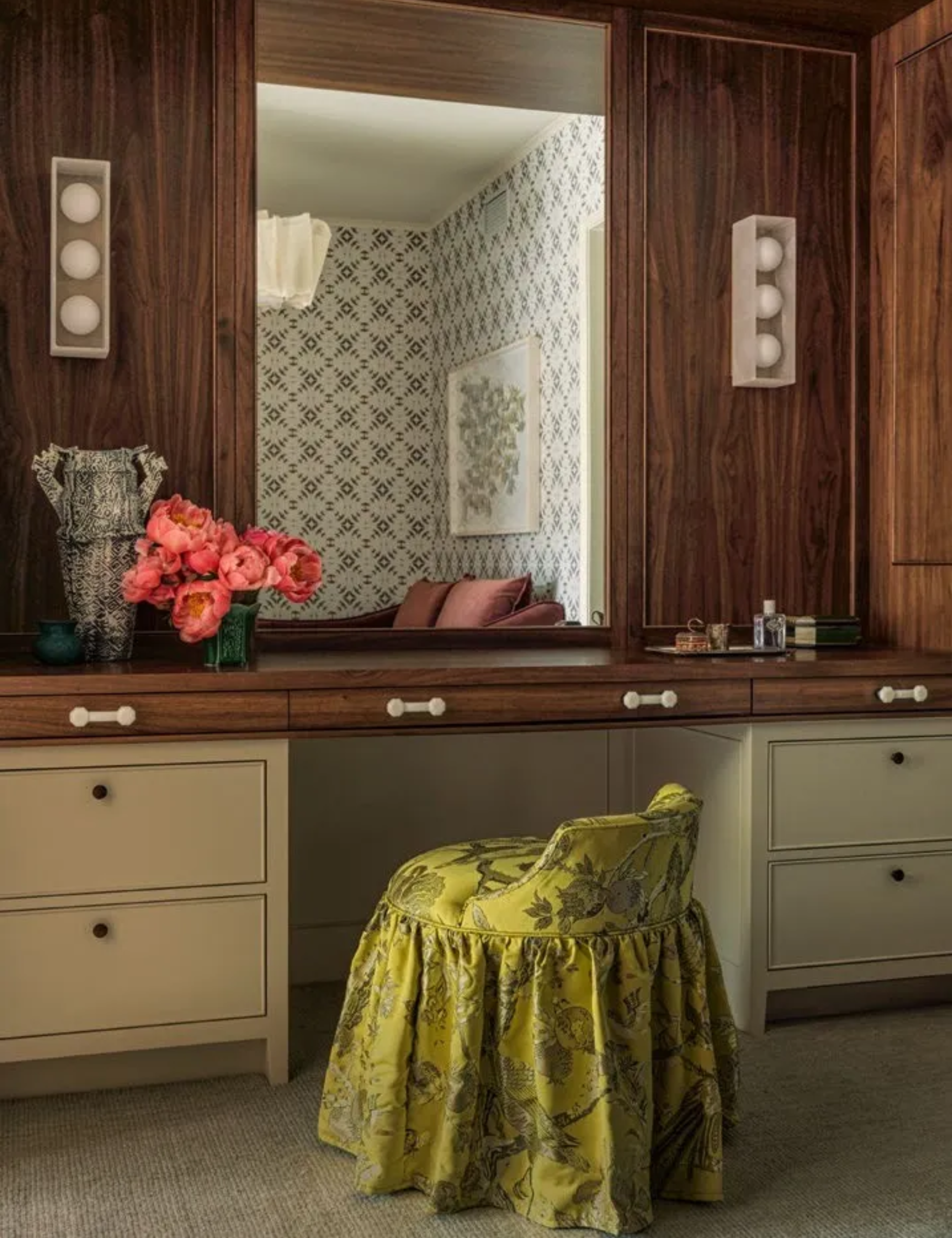 A stylish dressing room featuring a wooden vanity with a large mirror, a floral arrangement of pink flowers, and a vibrant yellow skirted chair against a patterned wall.