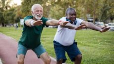 two older men performing a squat with arms outstretched in front of them in a park setting. 