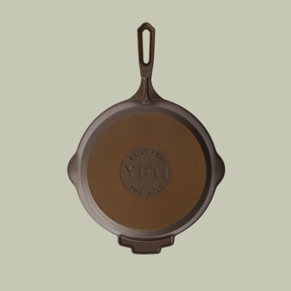 A cast-iron pan in brown with an inscribed &quot;YETI&quot; logo sits against a pale green background.
