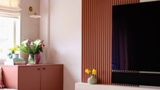 A living room with a tv on the wall. There is also a shelf under the tv with a vase and flowers. On the left side of the room are double door cabinets with books on top and a vase with flowers. 