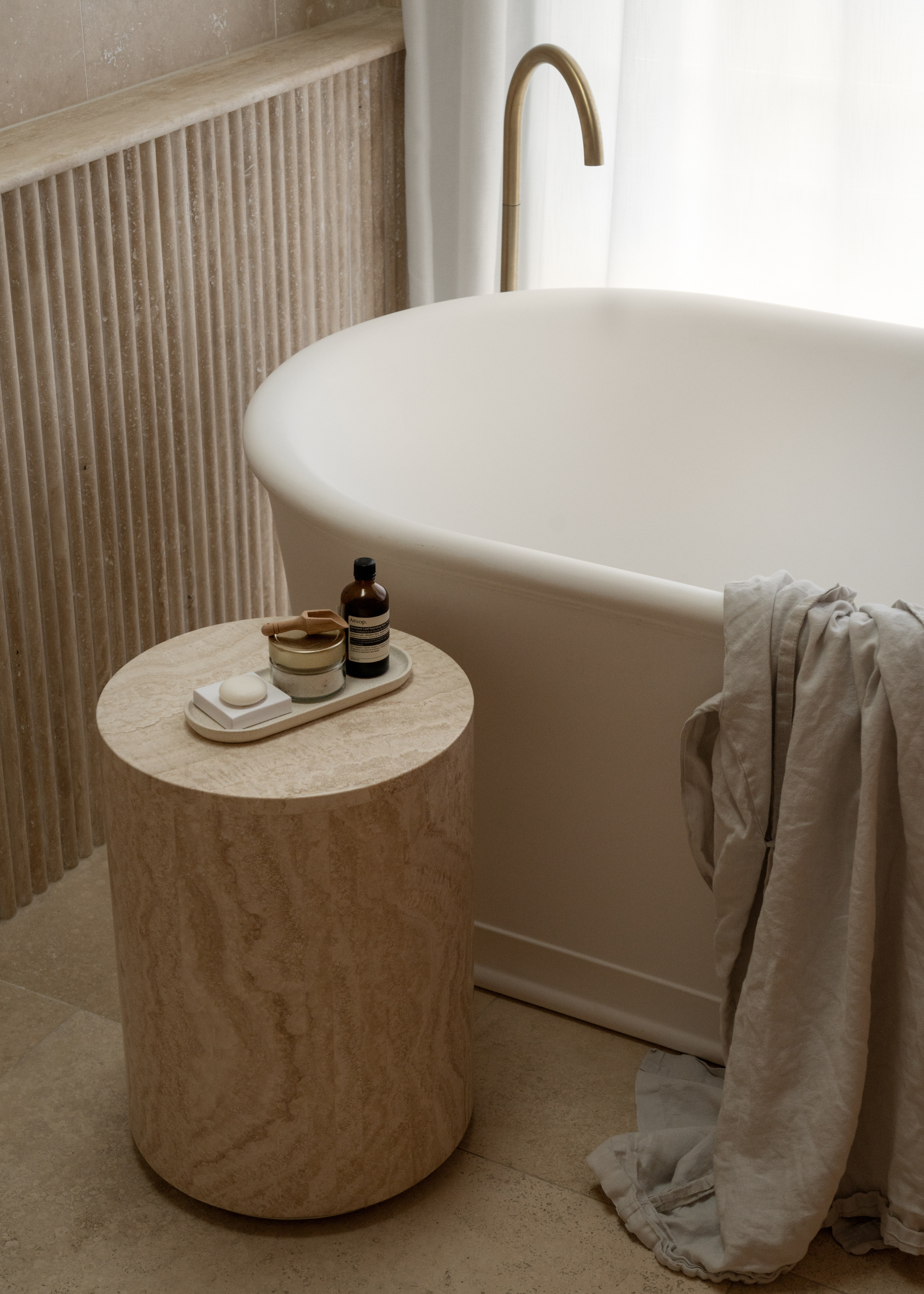 A minimalist bathroom with a bathtub with a draped shirt on the rim by a wood table with a tray of Aesop bath oils, bath salts, and a bar of soap