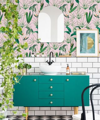 A bathroom with pink and green leafy wallpaper, a gold arched mirror, a dark green vanity with a white basin, a trellis plant to the left, and white and black checked flooring