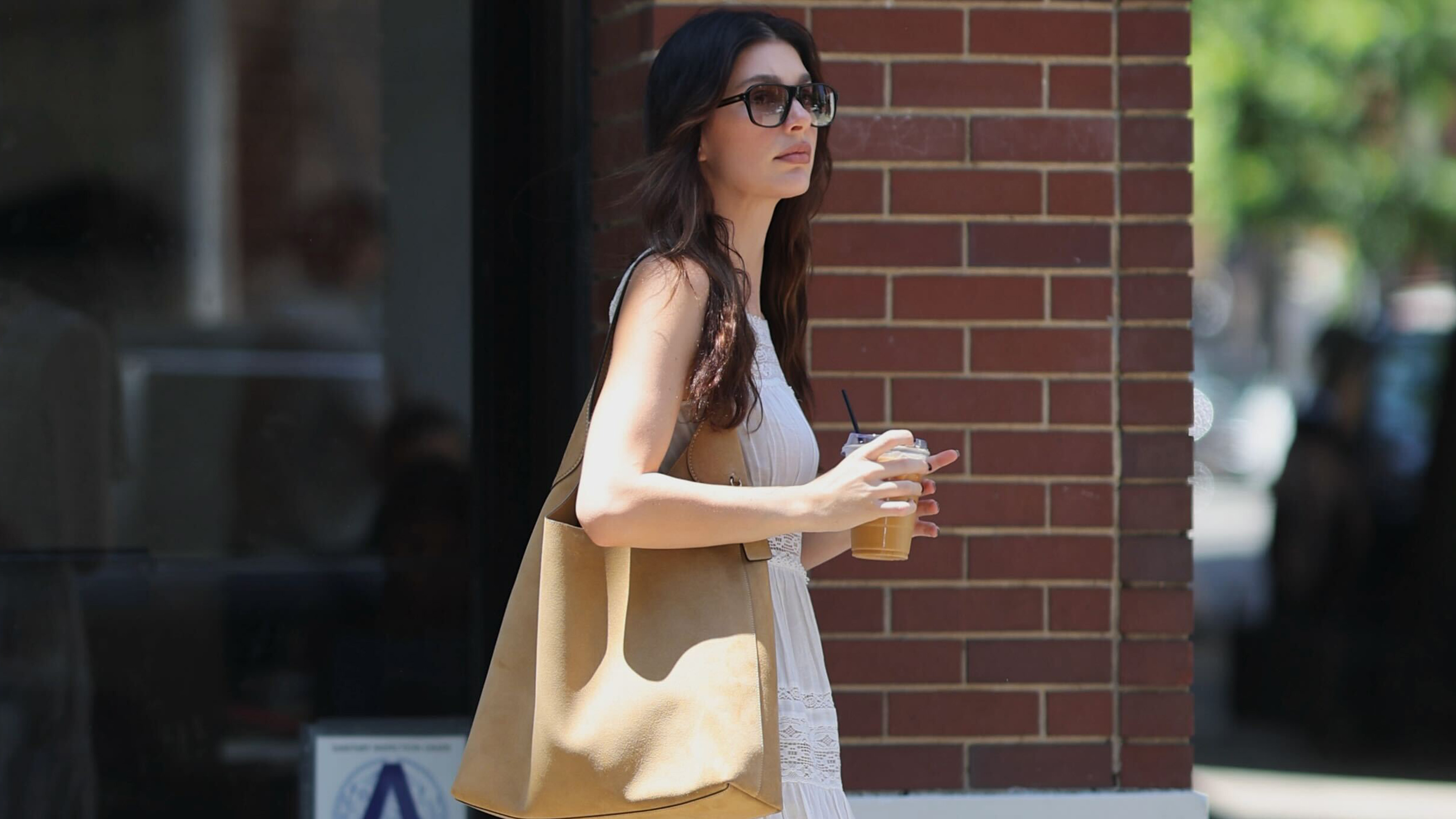Camila Morrone wears a white sundress, coach brooklyn bag, and cowboy boots.