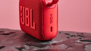 The USB C charging port on a red JBL Go 4 pictured against a pink background. It is sitting on a dark stone surface with puddles of water around it.