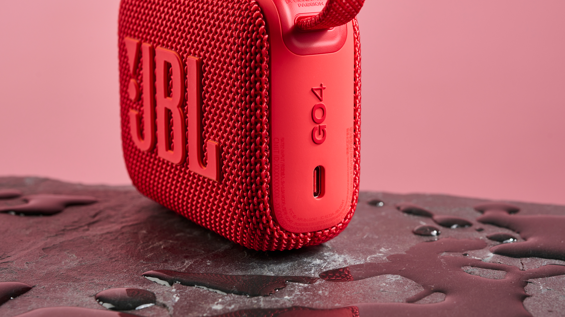 The USB C charging port on a red JBL Go 4 pictured against a pink background. It is sitting on a dark stone surface with puddles of water around it.