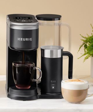 A Keurig K-Cafe coffee maker on a white countertop against a beige wall; to the right, a cup of coffee in a glass mug.