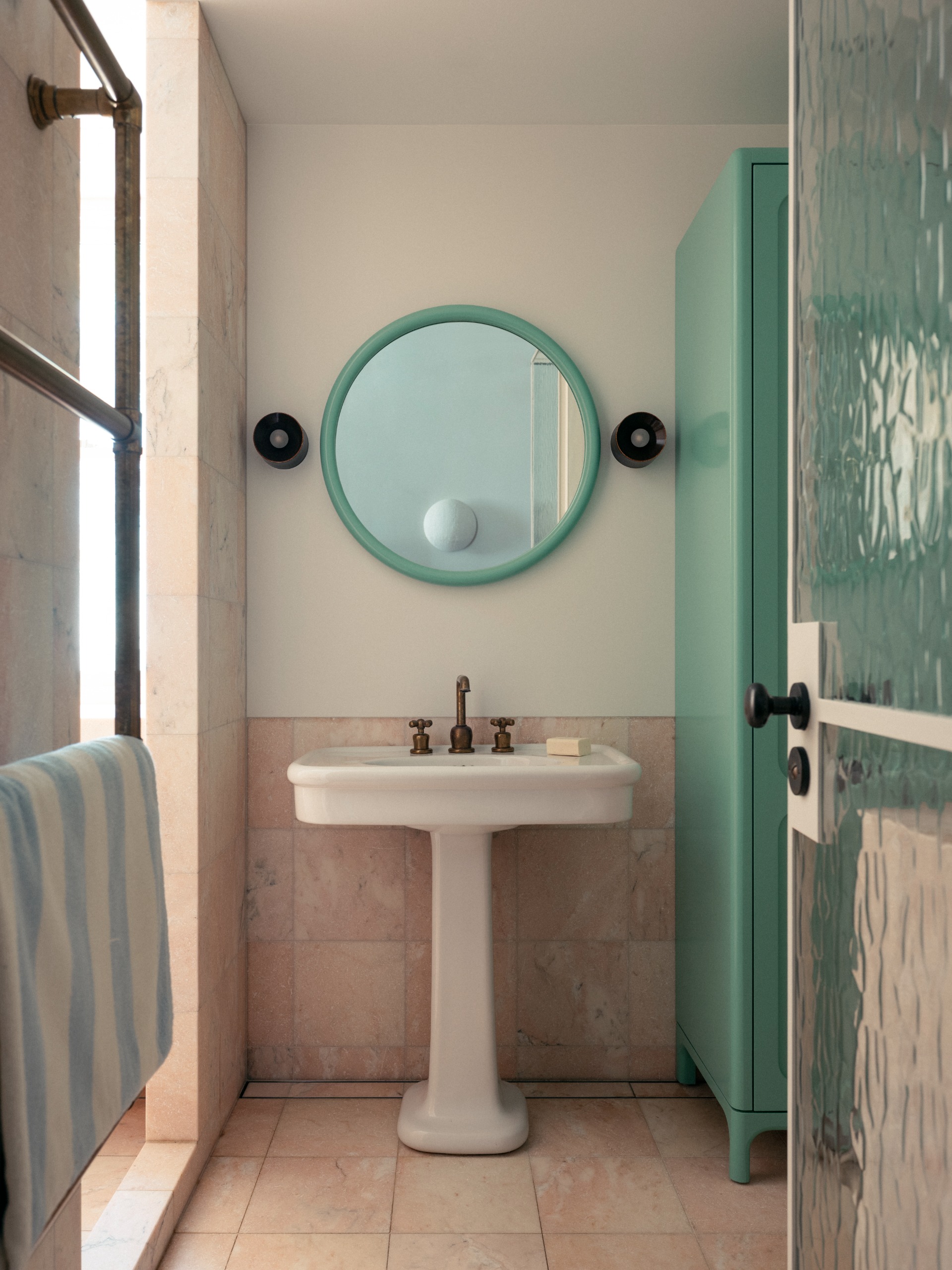 bathroom with mint green round mirror, white pedestal basin, cream tiles halfway up the walls, mint green cabinet, and textured glass door with striped towel