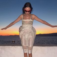 Alexa Chung wears a white lace-trim knee-length dress with white ballet flats and red sunglasses.