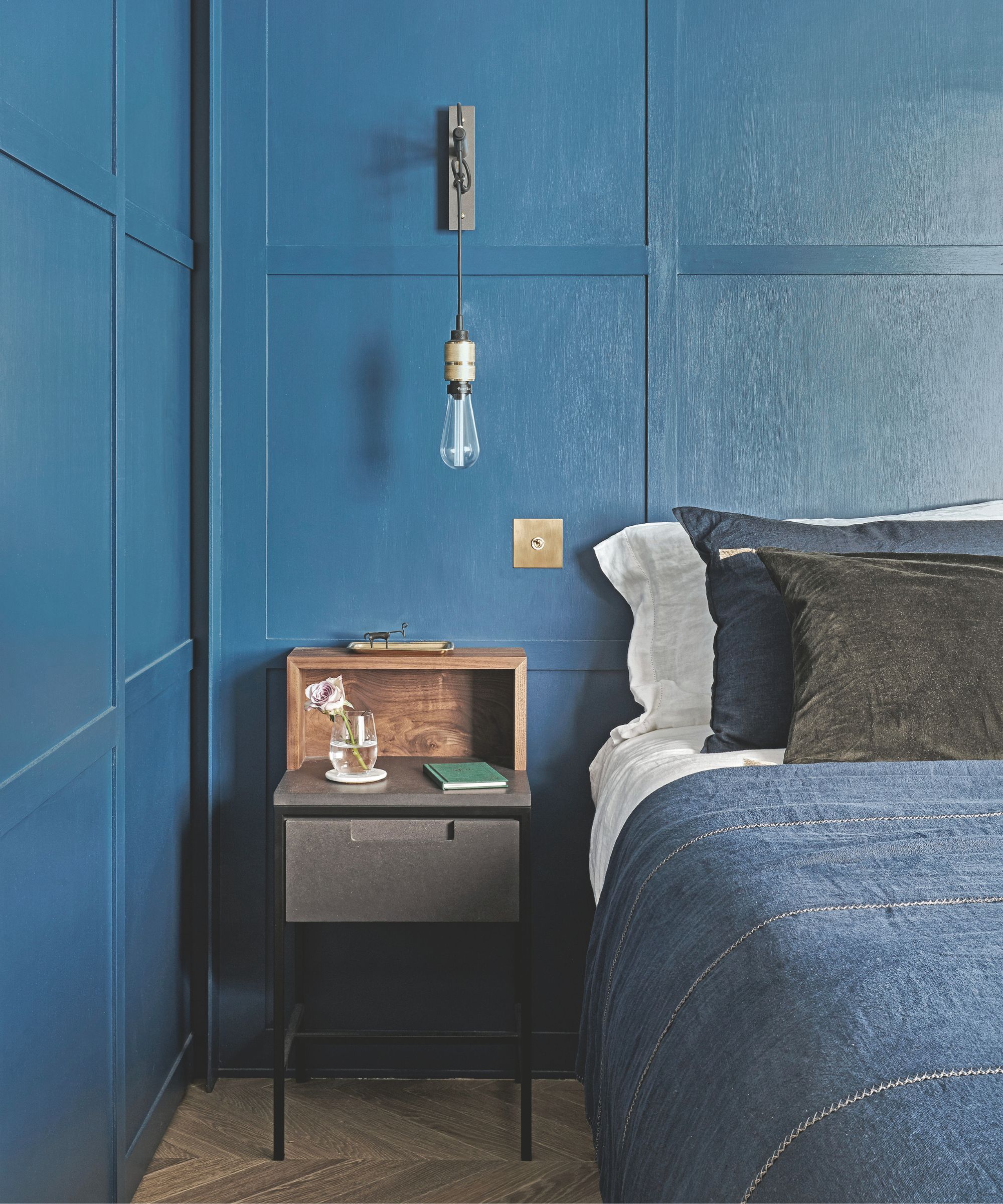 A blue bedroom with blue panelled walls, a bed with white bedding, blue blankets, and blue and brown cushions and pillows. To the left of the bed is a bedside table with a book and flower, and a sconce lamp is hanging above.