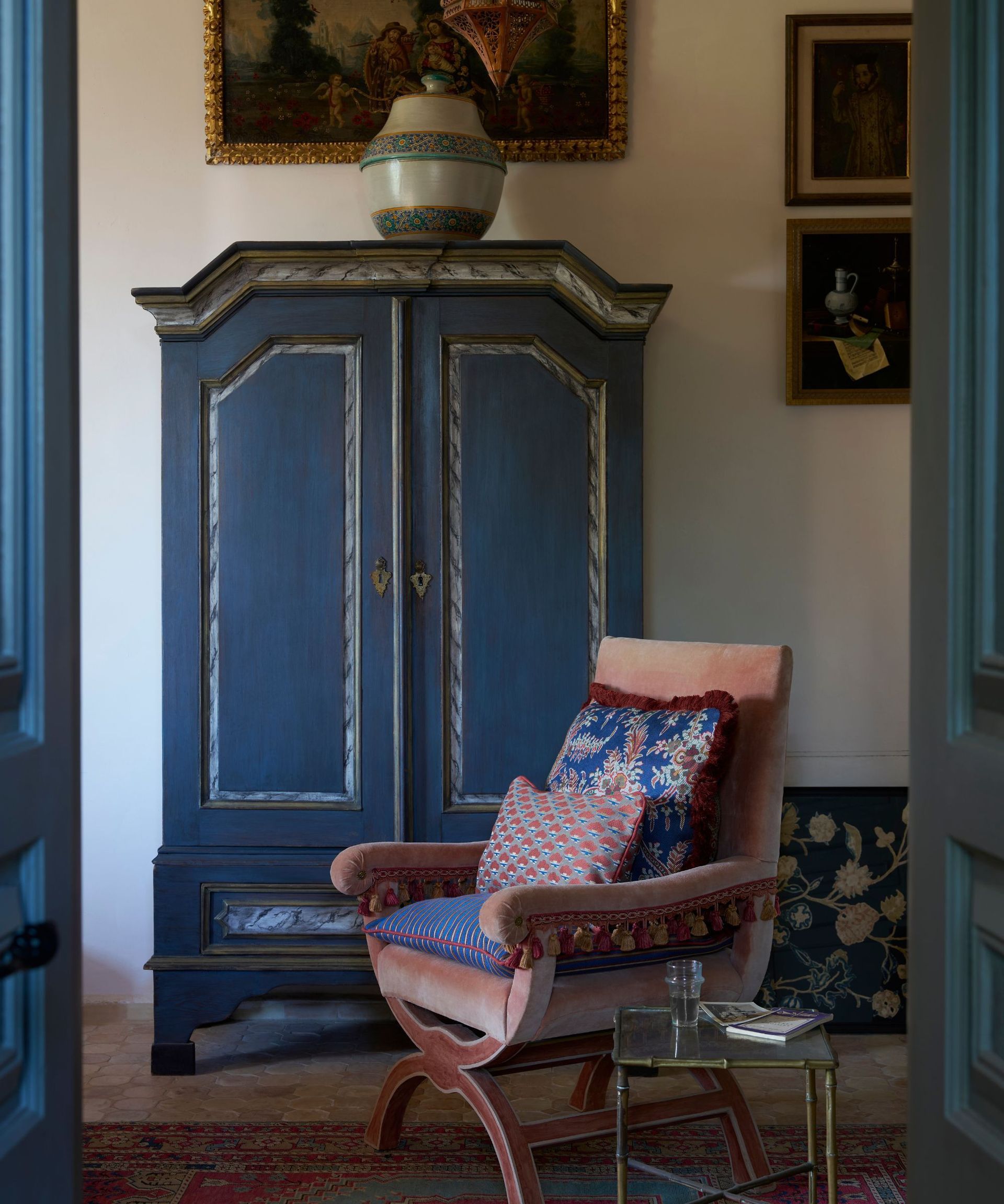 A bedroom with a large blue wardrobe and a salmon pink velvet chair, with cushions upholstered in Liberty fabric