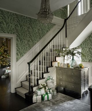 Christmas stair decor ideas with green wallpaper, green and silver presents at the foot of the stairs, and pine branches in a vase on the table