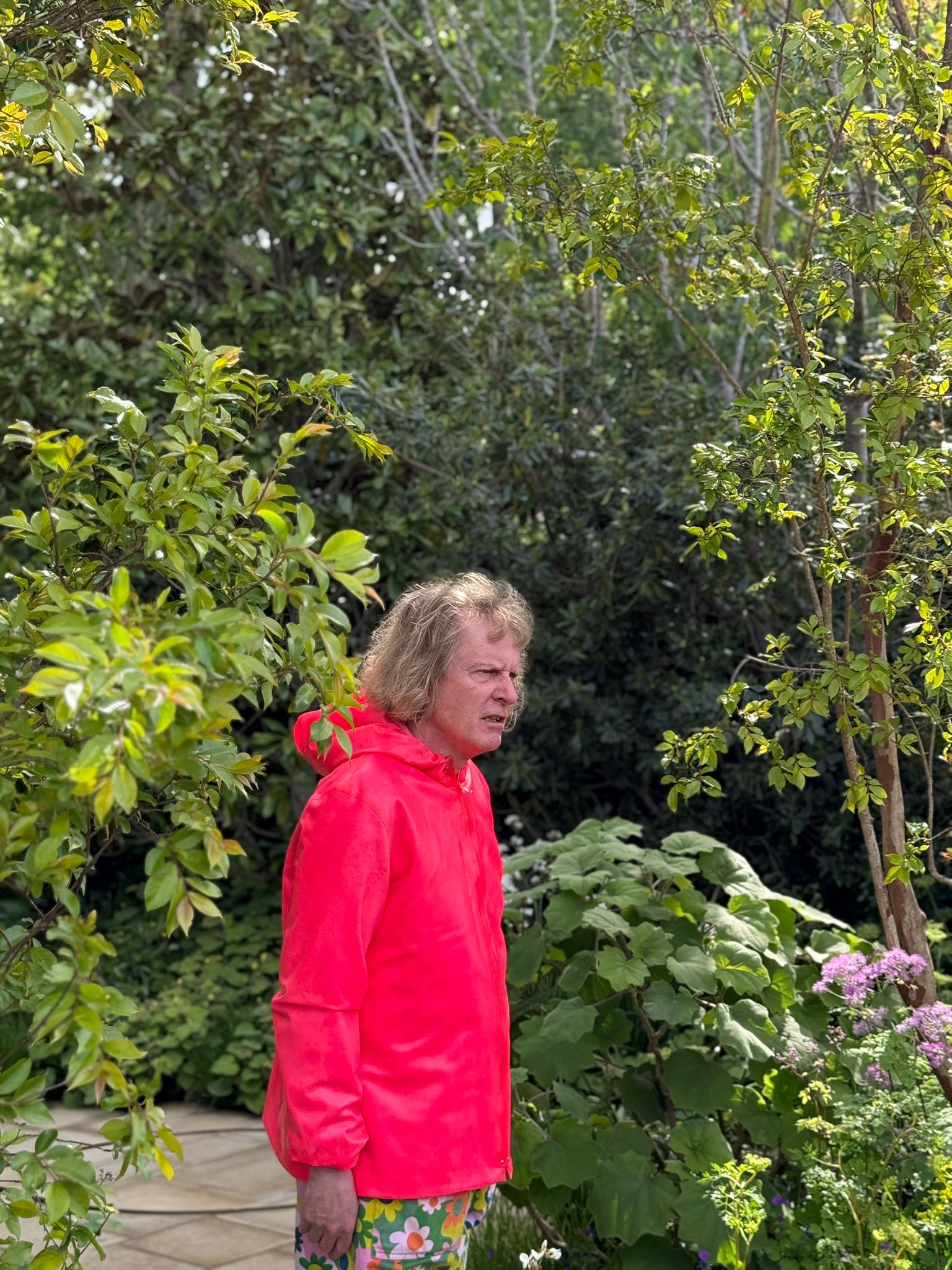 Grayson perry, in a red anorak, walks among the plants and trees of the Tate Britain Garden