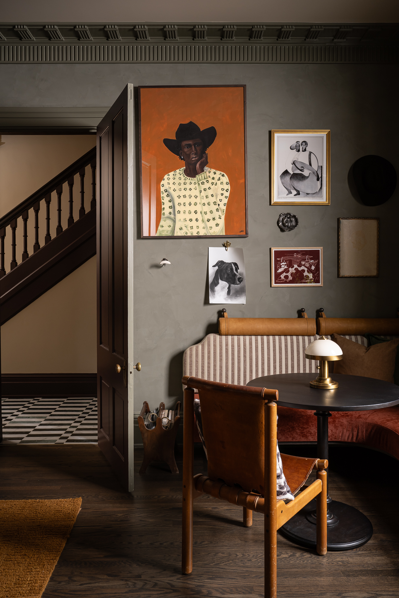 Grey washed walls with decorative wall art including a man on a orange background with cowboy hat and white patterned jumper and a sketch of a dog, with a round black table, lamp and chair