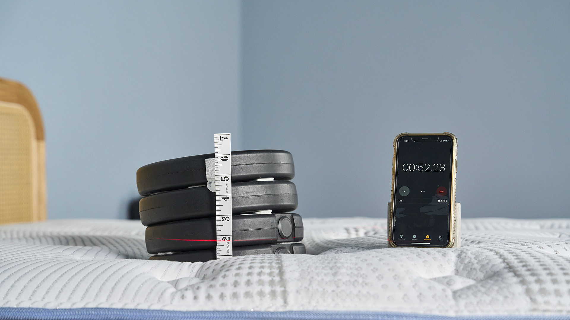 A close-up shot of the white top of the Bear Elite Hybrid mattress with a stack of four black, circular weights resting on it. A measuring tape is stuck vertically against the stack, showing a compression of about 2.5 inches with a red laser line marking that measurement. A smartphone next to the weights displays a stopwatch reading 00:52.23.