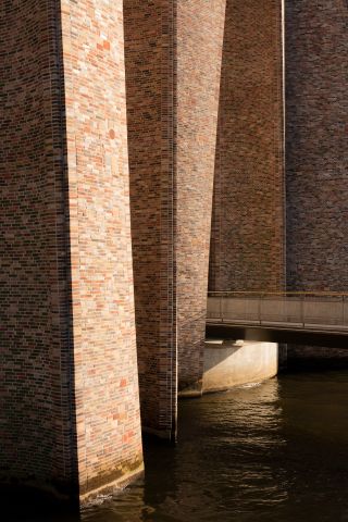 Fjordenhus, by Olafur Eliasson and Studio Olafur Eliasson in Vejle, Denmark. Large brick walls with a bridge going between them over a body of water.