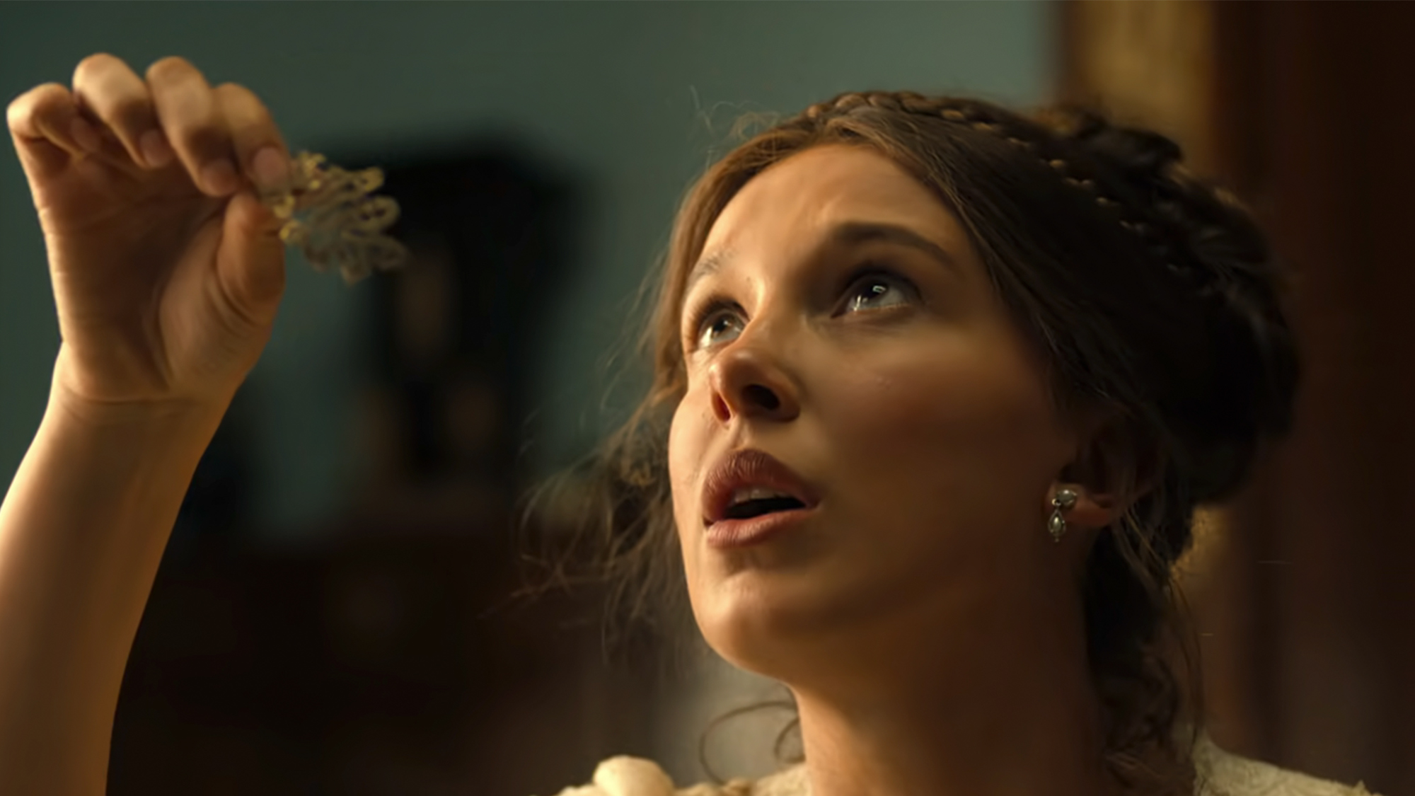 Millie Bobby Brown looking up and holding something in her hand in Enola Homes 3