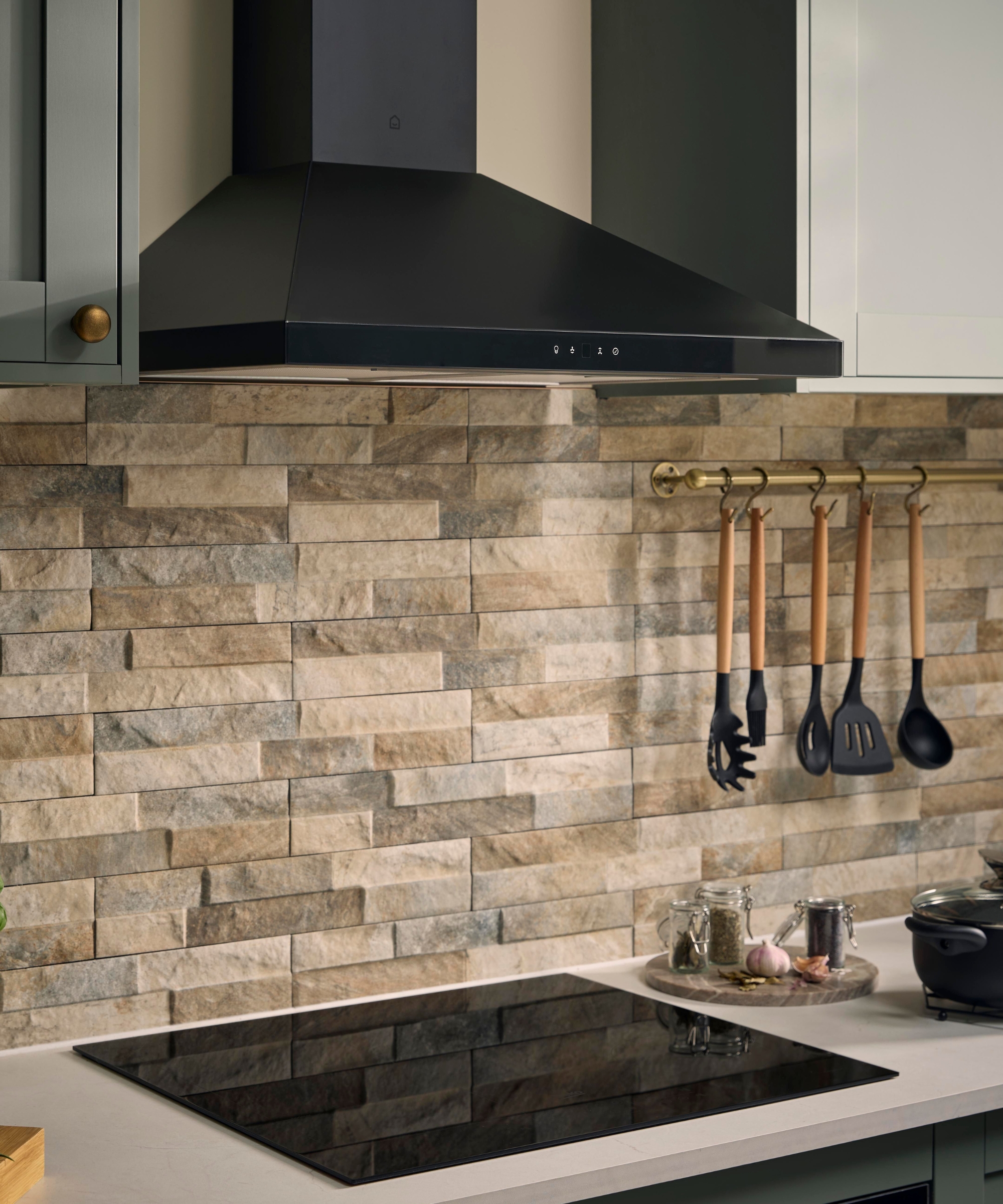 Black extractor hood above an induction hob, with a stone-tile splashback and hanging utensils.