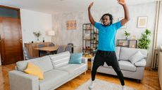 A man in shorts, leggings and a t-shirt works out in a living room. He is performing a star jump, with his legs out wide and his arms overhead. Behind him we see a couch, a dining table and decorative shelving.