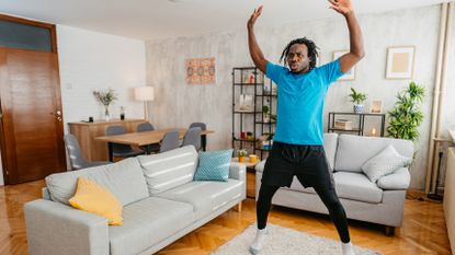A man in shorts, leggings and a t-shirt works out in a living room. He is performing a star jump, with his legs out wide and his arms overhead. Behind him we see a couch, a dining table and decorative shelving.