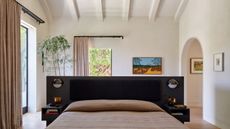 Image of a light, white, and open bedroom with a bed with a black head board that is floating in the room. There are two large windows with double curtain rods.