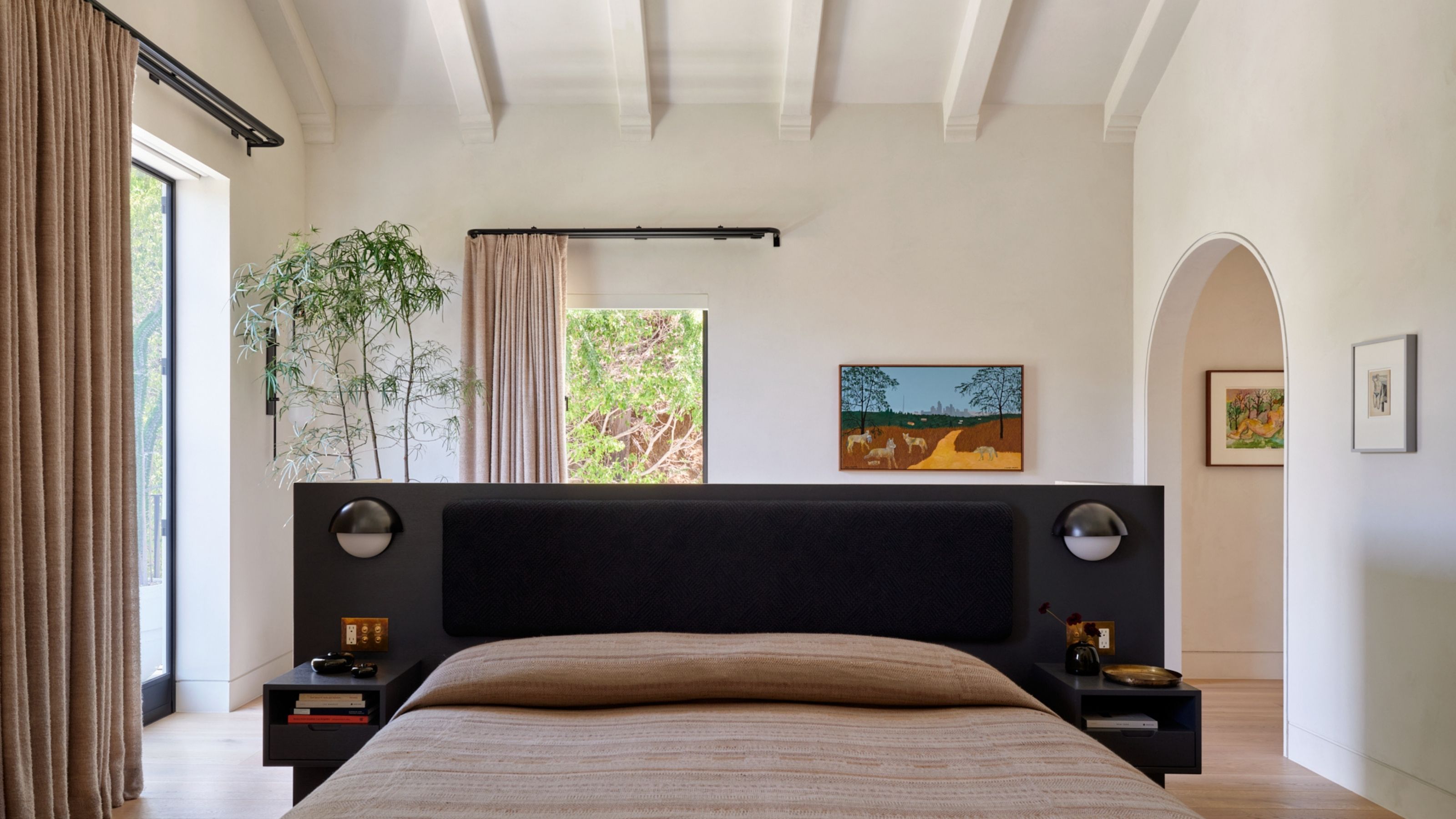 Image of a light, white, and open bedroom with a bed with a black head board that is floating in the room. There are two large windows with double curtain rods.