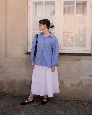 Editor Halie LeSavage wears a blue button-down shirt and white skirt with Miu Miu ballerinas and a blue hair tie