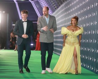 Robert Irwin in a tuxedo, Prince William wearing a gray suit coat and sneakers and Nomzamo Mbatha wearing a yellow gown with a slit while walking the Earthshot Prize 2024 green carpet.