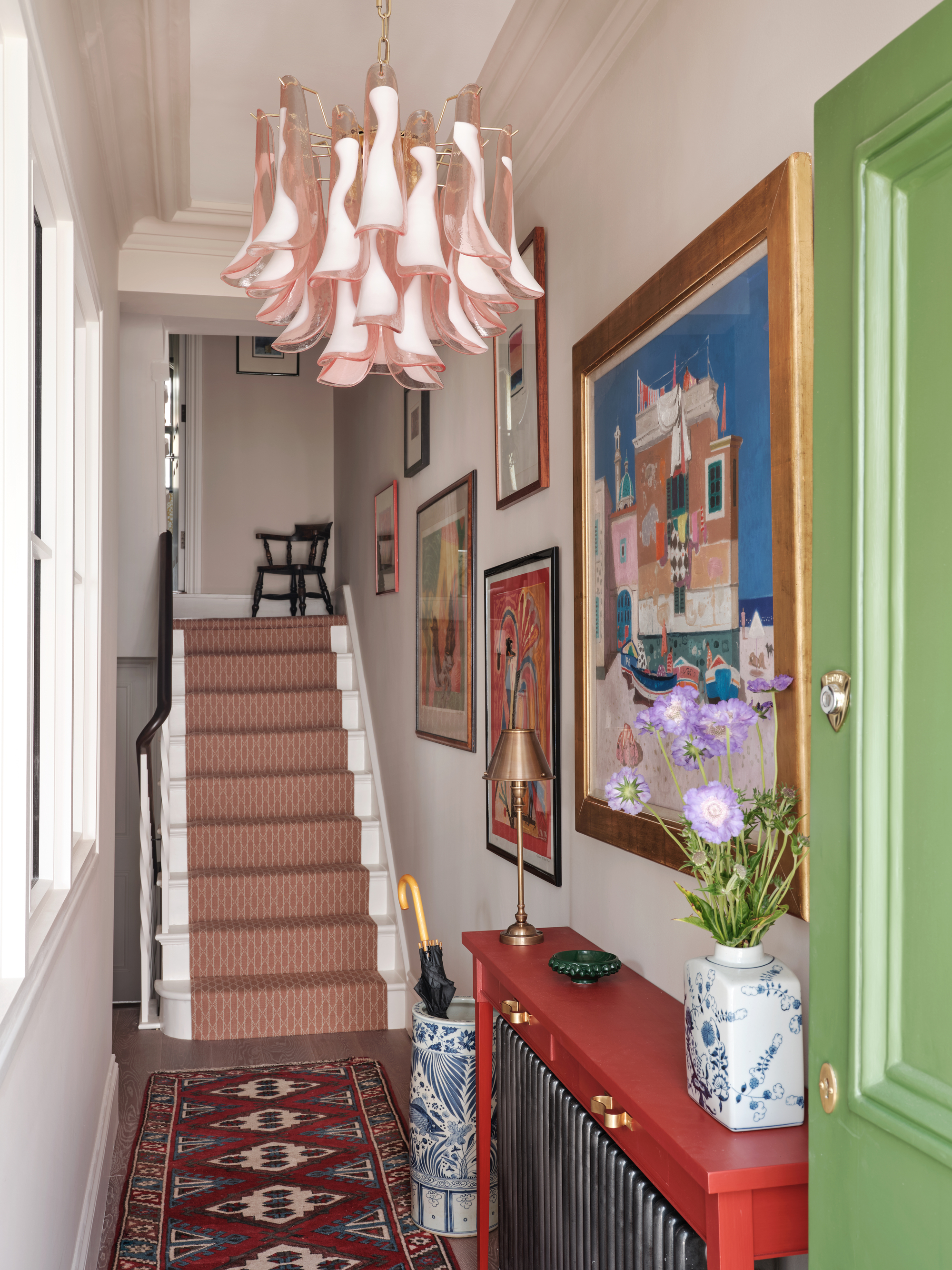 home entryway with gallery wall of artwork, a red console table over a radiator, and entryway rug with a pattern, a red patterned stair runner, and a sculptural pink glass pendant light