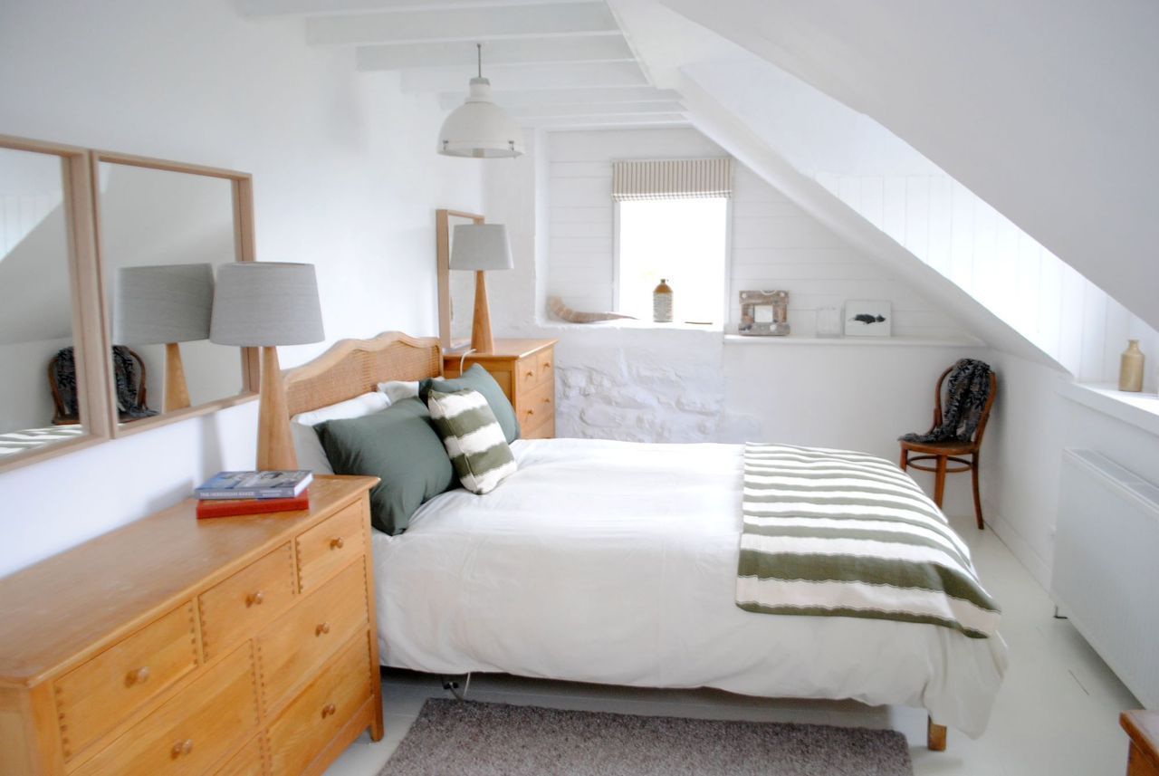 The white bed linen is surrounded by white stone walls which have two wooden framed mirrors on them and radiator and window opposite the bed