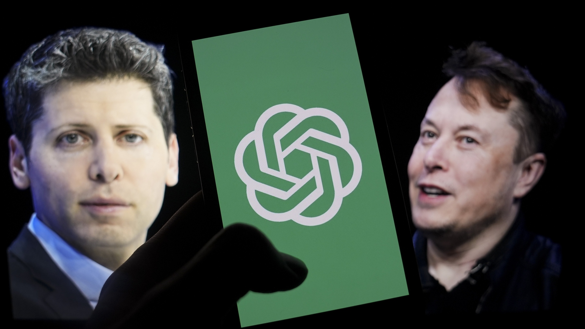 The logo of 'OpenAI' is displayed on a mobile phone screen in front of a computer screen displaying the photographs of Elon Musk and Sam Altman.
