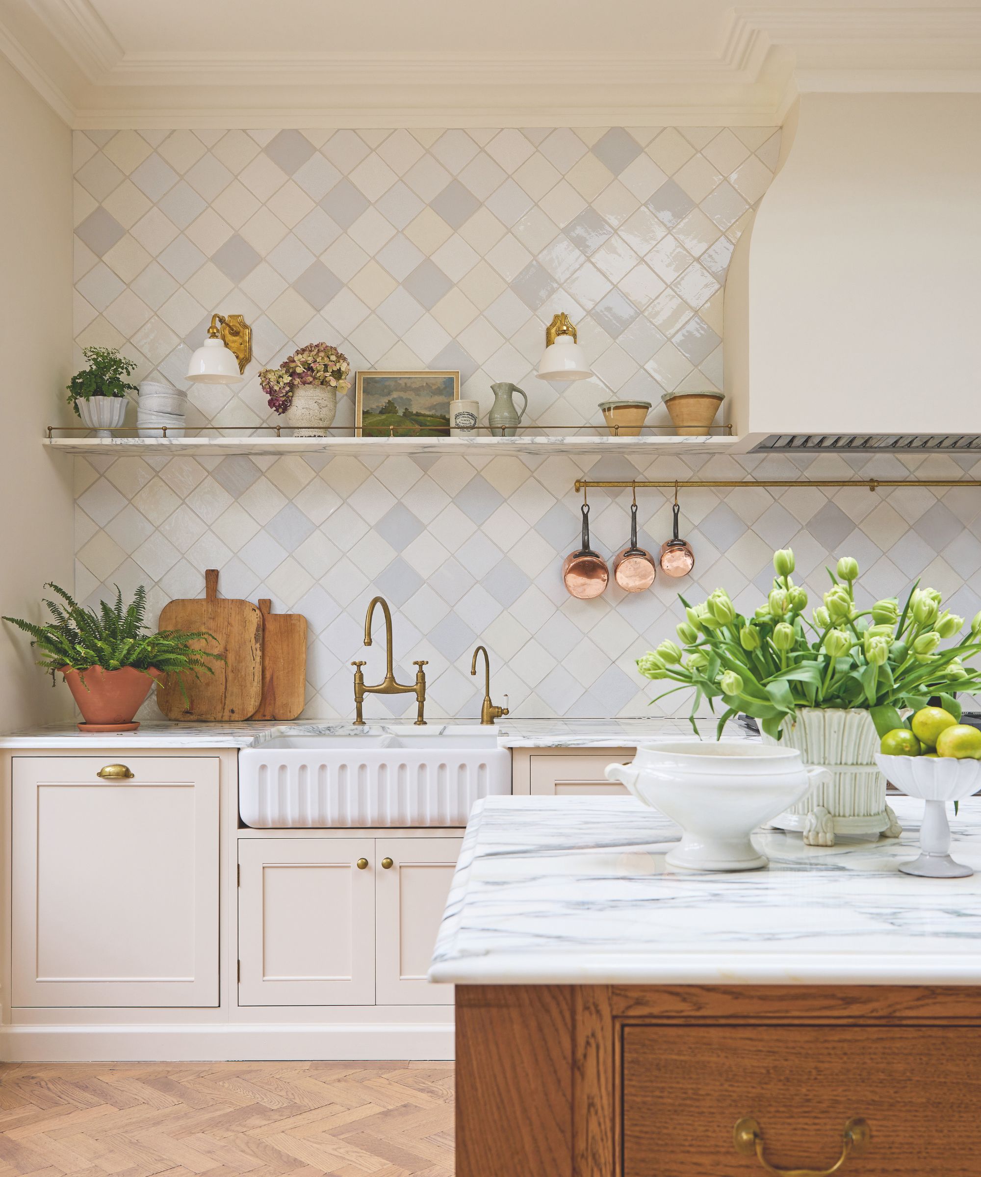 Close-up of a kitchen workspace featuring a white ceramic apron-front sink and antique brass faucets. Above, a marble floating shelf displays curated ceramics and small paintings against a backdrop of glossy diagonal tiles