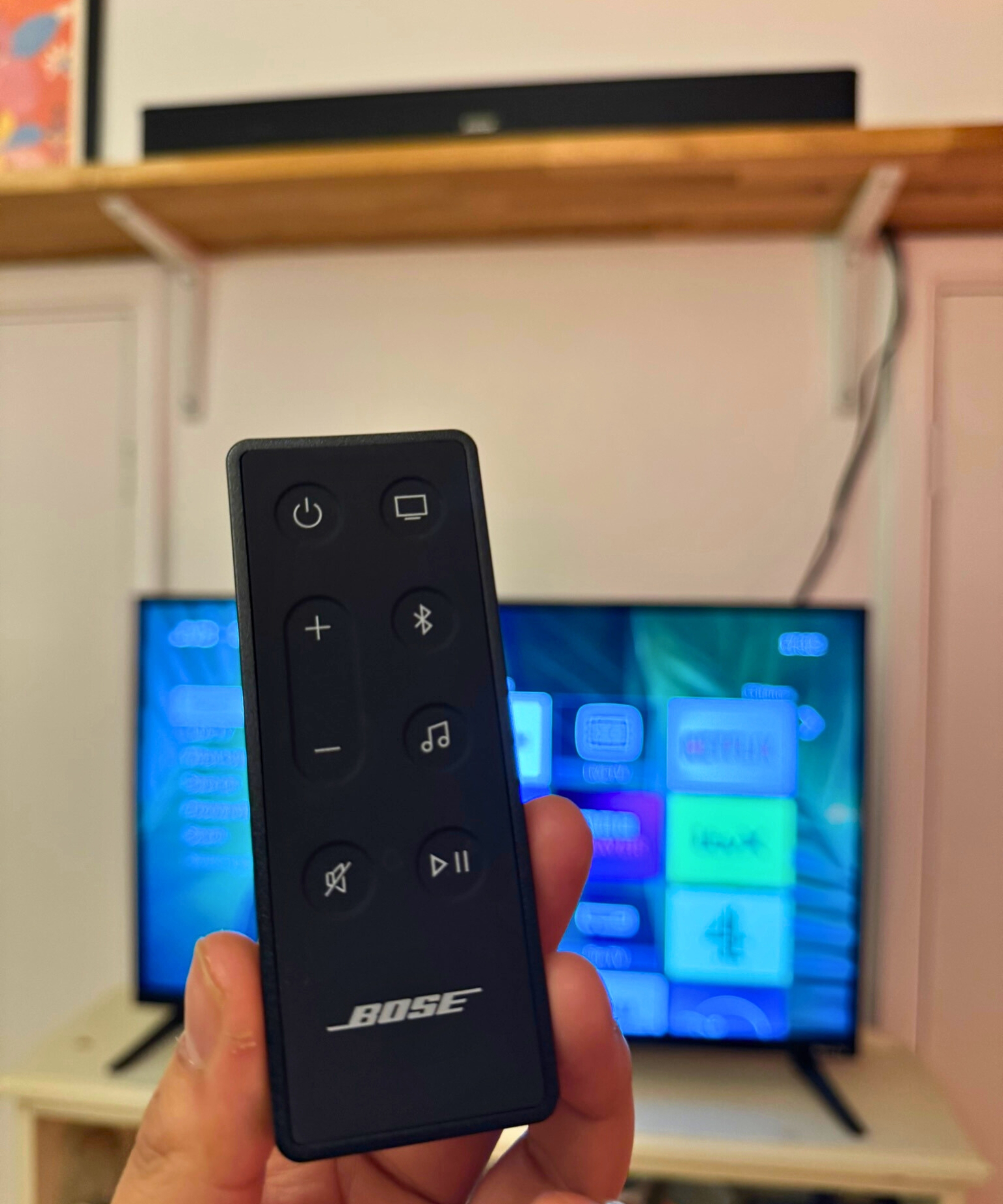 Hand holding the remote for the Bose Smart Soundbar in front of TV, soundbar, and shelf.