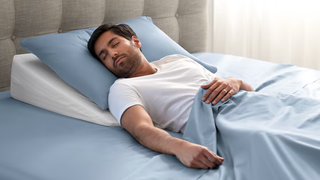A man sleeping on a slanted white pillow in bed with an extra blue pillow and a blue blanket on top