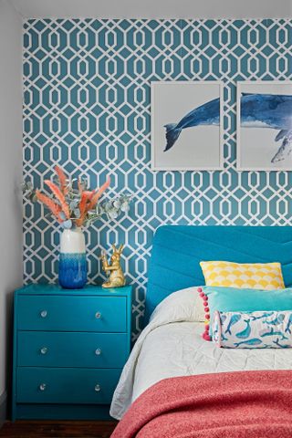 Guest bedroom with blue upholstered bed against blue geometric feature wallpaper, blue bedside chest of drawers and pink throw