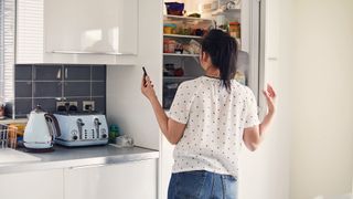 Woman with dark brown hair wearing a white top looking into an oven white fridge.