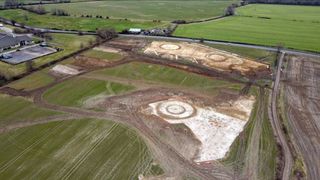 An aerial view of the site, which includes dirt fields with round barrows within a larger green field in the countryside.