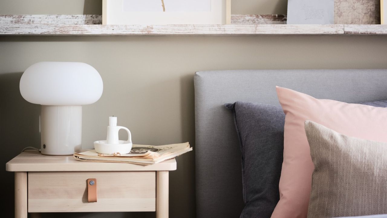 IKEA DEJSA table lamp in a calm, neutral bedroom with a wooden bedside table, bed, and floating shelves with framed art