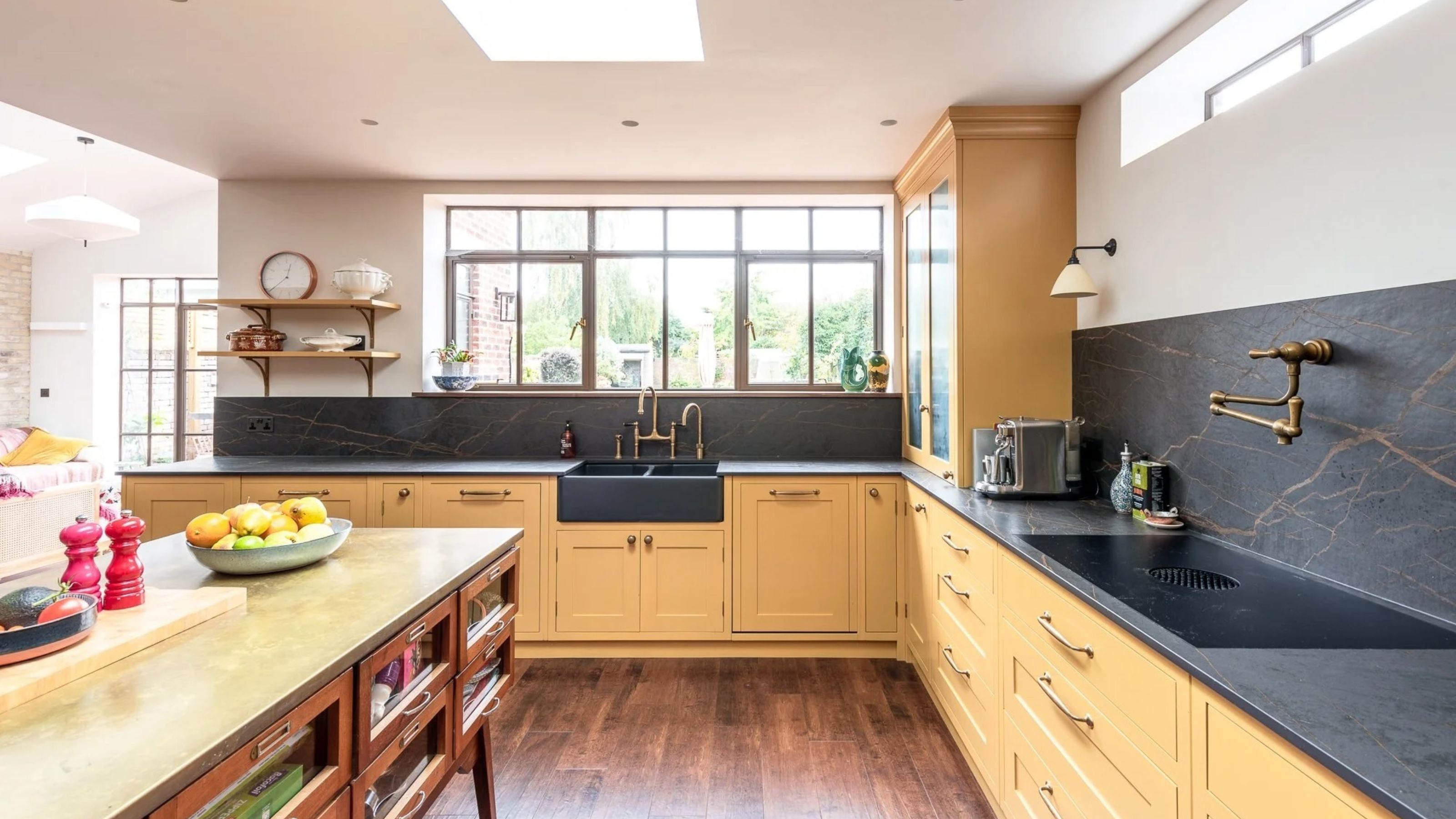 yellow kitchen with timber floors and black stone countertop and backsplash with brass taps and pot filler, and timber table used as a kitchen island bench