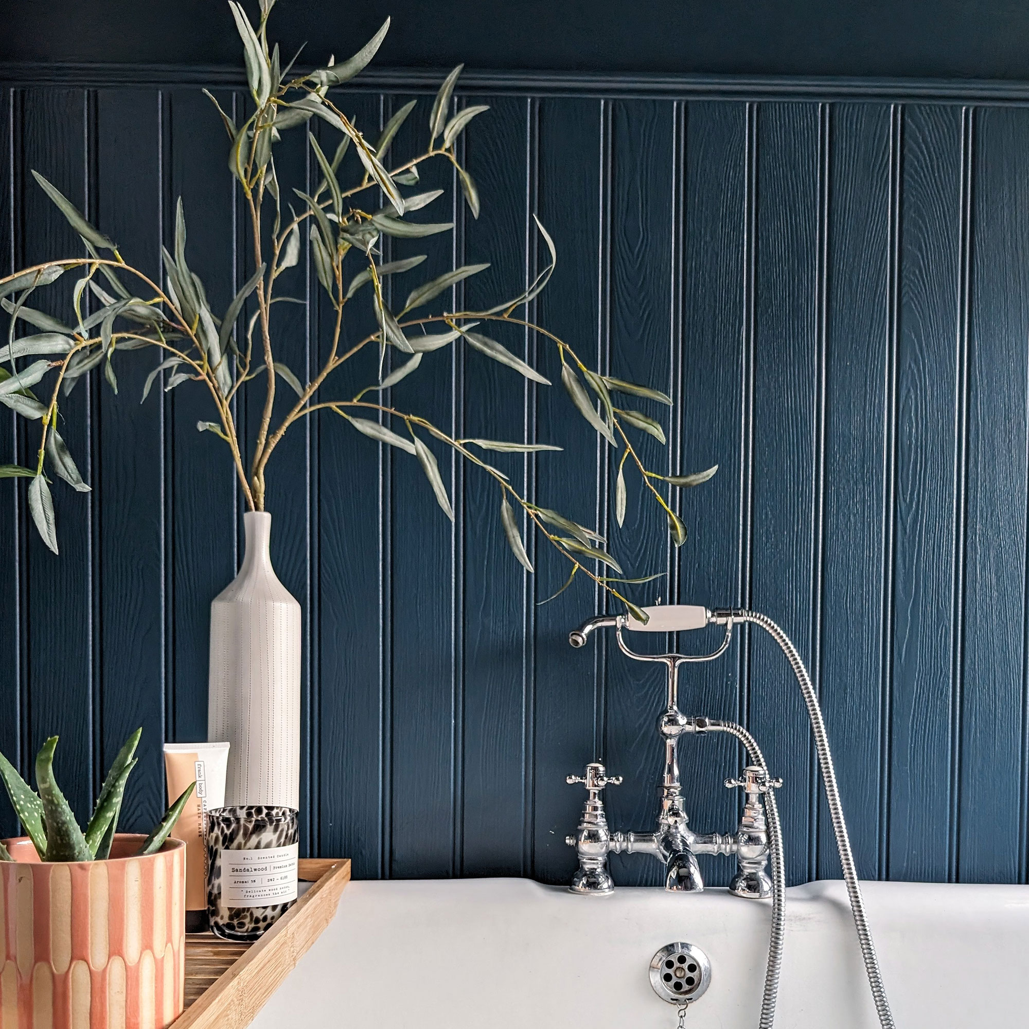 Wooden bath tray on bath holding plant, candle, lotion and vase in bathroom with teal panelled walls