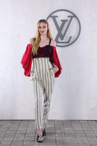 Sophie Turner at Louis Vuitton's PFW show wearing striped suspender pant with a red draped blousents with a