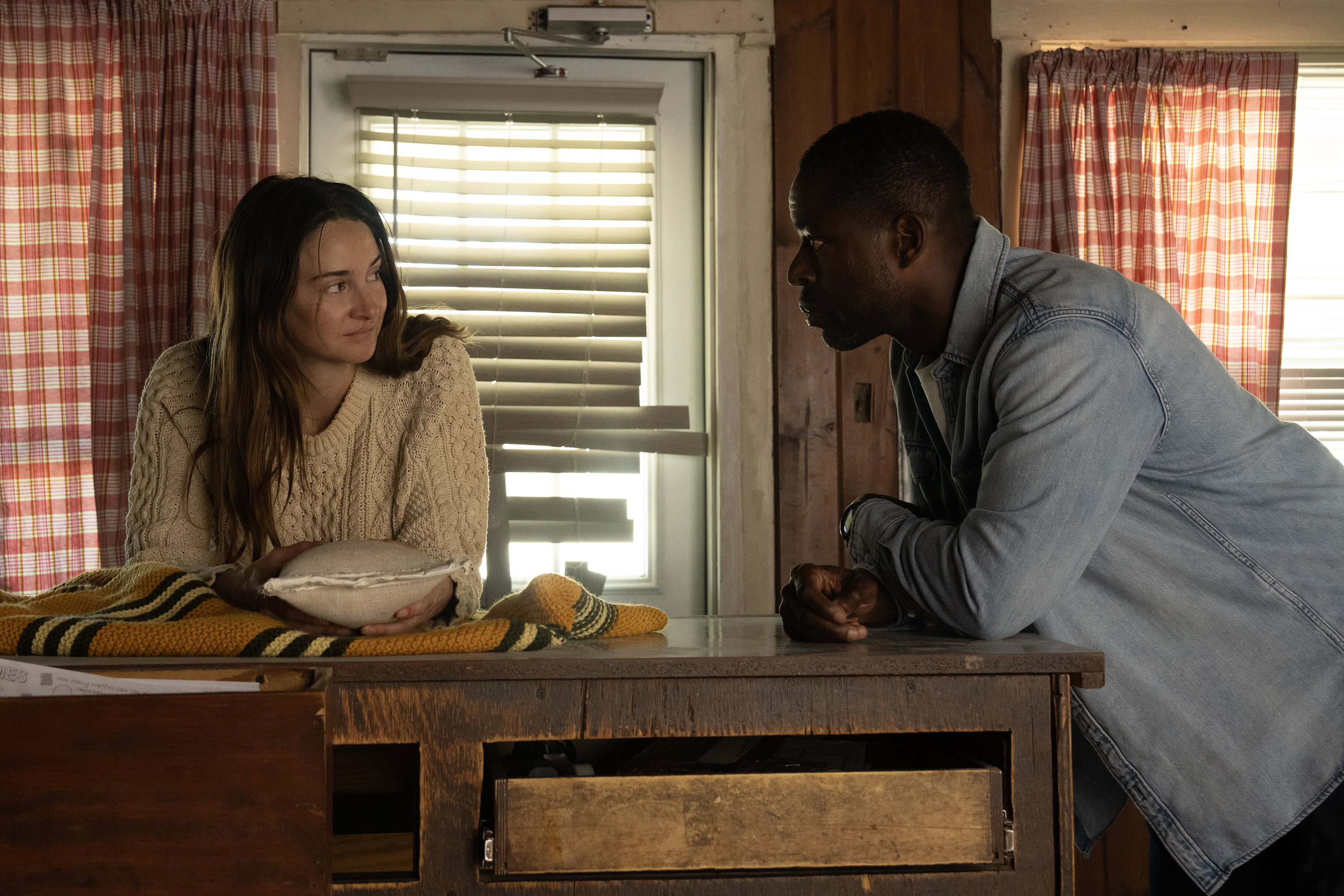 A woman in a white sweater (Shailene Woodley) and a man in a denim shirt (Sterling K. Brown) lean on a wooden counter in an abandoned diner. A stuffed bag sits on a yellow striped blanket on the counter, in a still from 'Paradise' season 2.