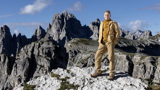 Chris Packham in a mustard jacket and trousers stands in front of the Dolomite mountains in Earth documentary.